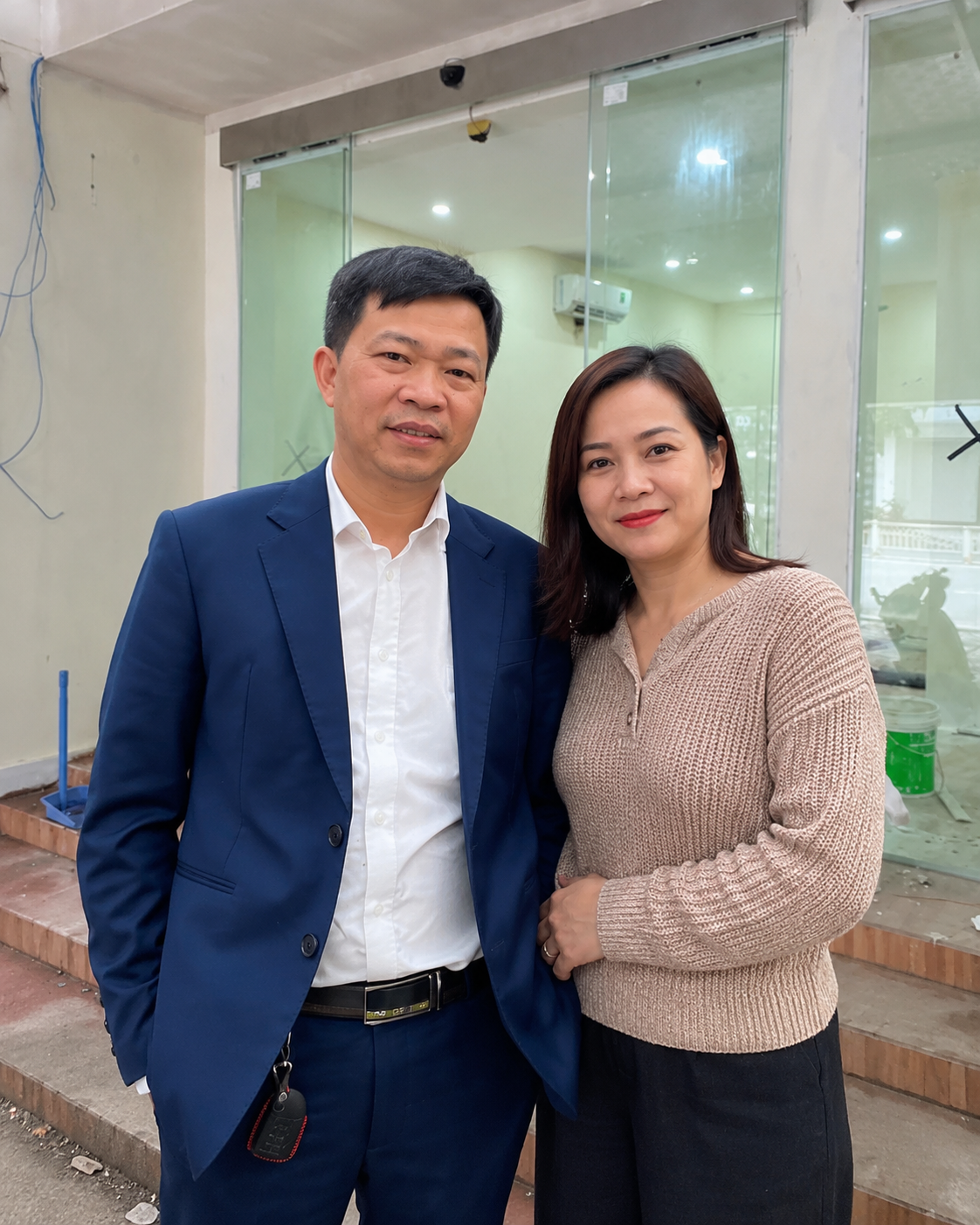 Hai người mặc vest xanh và áo len be đứng trước cửa kính công trình đang hoàn thiện